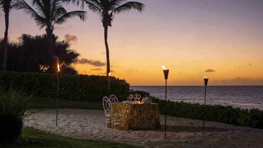 Private dining table with gold tablecloth, torches on sand beach at sunset