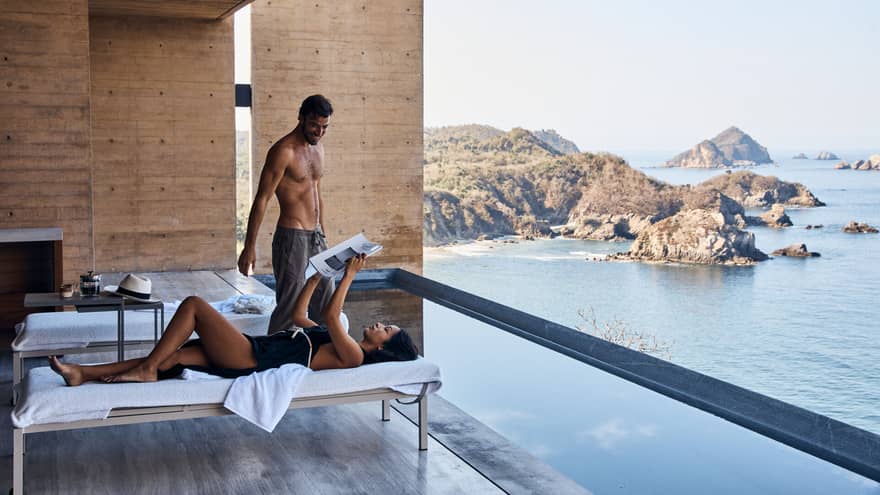 A couple unwinding on a luxurious lounge chair, reading a magazine and chatting, on a balcony overlooking a shimmering ocean and beautiful cliffs