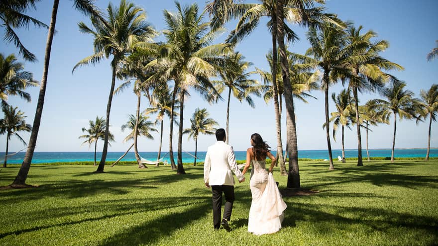 Back of bride and groom running across tropical lawn with palm trees towards ocean