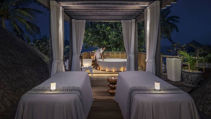 Outdoor night spa setting with two massage tables, soft candlelight, and a staff member preparing a bathtub under a canopy surrounded by tropical greenery