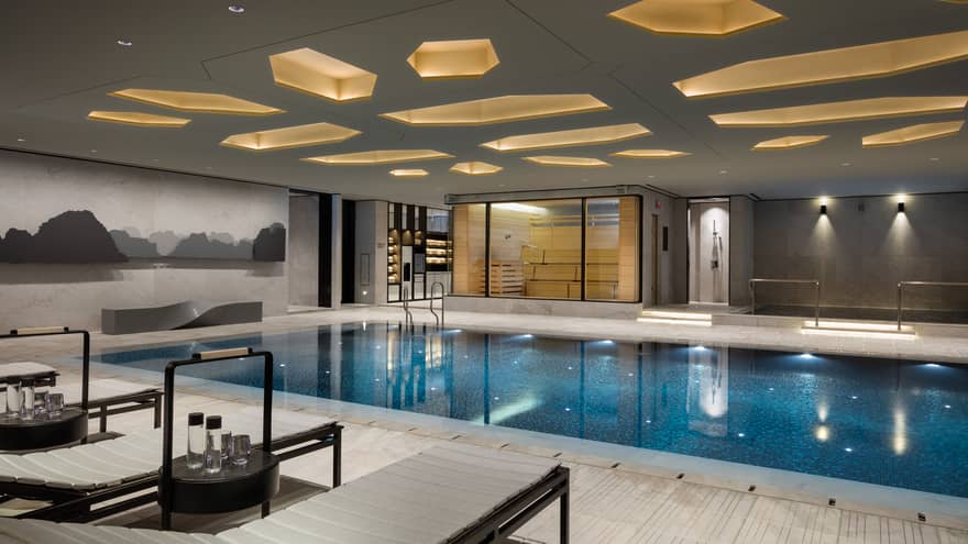 Lounge chairs, tables with water on deck by indoor swimming pool under ceiling with decorative recessed lights