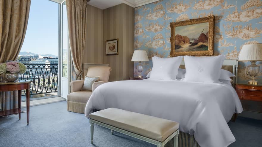 Luxury hotel room with a king bed, nightstands with lamps, bench, patterned wallpaper, armchair, framed artwork and a balcony with city view