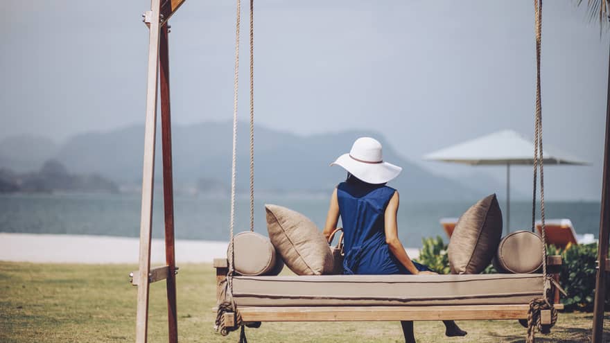 Woman in blue dress and white sun hat lounges on outdoor swing