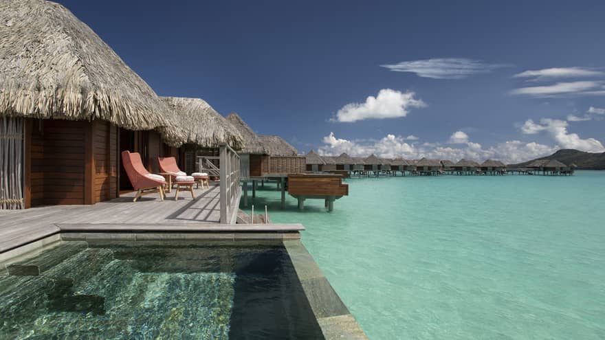 Overwater bungalows with thatched roofs, private pools and lounge chairs overlooking a clear turquoise lagoon under a bright blue sky.