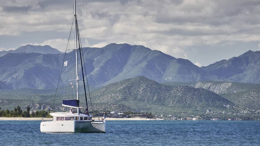 A catamaran on calm, blue water under a partly-cloudy sky with low green hills in the background and mountain ranges beyond.