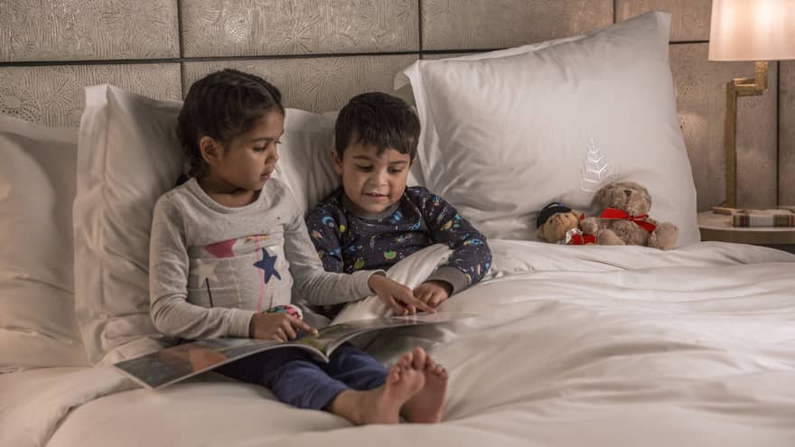 Young girl reads to her brother at bedtime, both in pajamas, a teddy bear propped on the pillow next to them