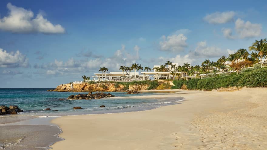 View across white sand beach to modern white event venue on rocks over sea 