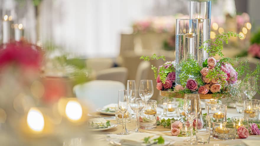 A wedding table setting featuring soft flickering candles in long glass holders, pink roses centerpiece and champagne glasses