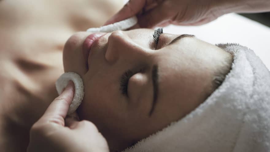 A detail of hands rubbing a sponge on a woman's face as she lies on a massage table with her eyes closed and hair wrapped up in a white towel