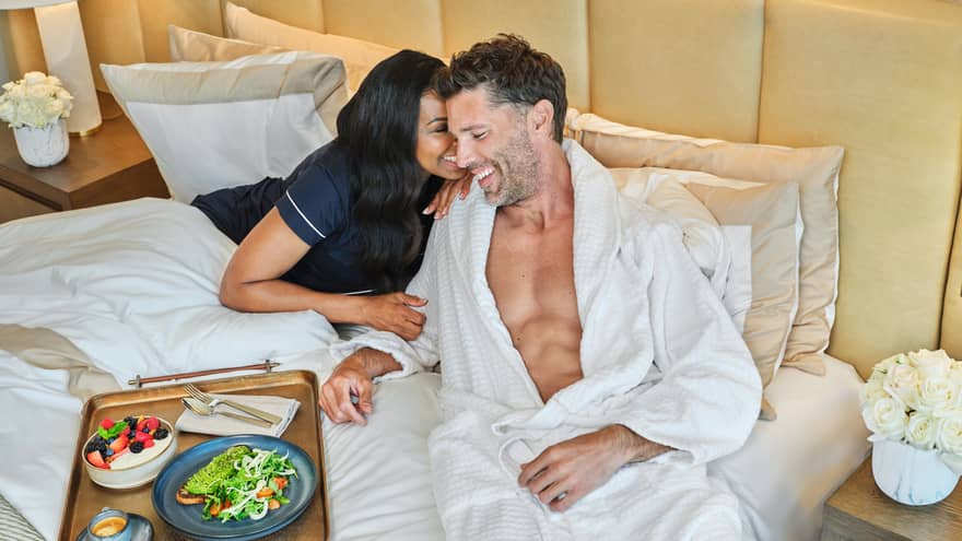 A couple cuddles in bed smiling with a tray of breakfast on the bed next to them