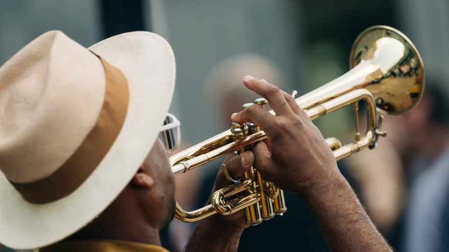 A man wearing a cream-and-taupe fedora with a brown ribbon plays a trumpet