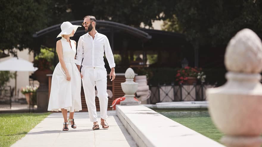A couple dressed in white cotton attire strolls leisurely beside a peaceful pond, enjoying the sunny garden ambiance