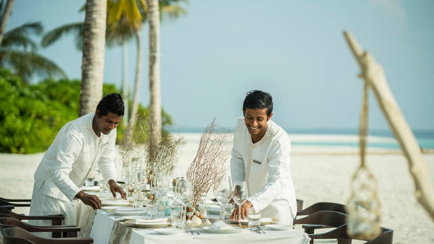 Two hotel staff set plates, wine glasses on private dining table on white sand beach