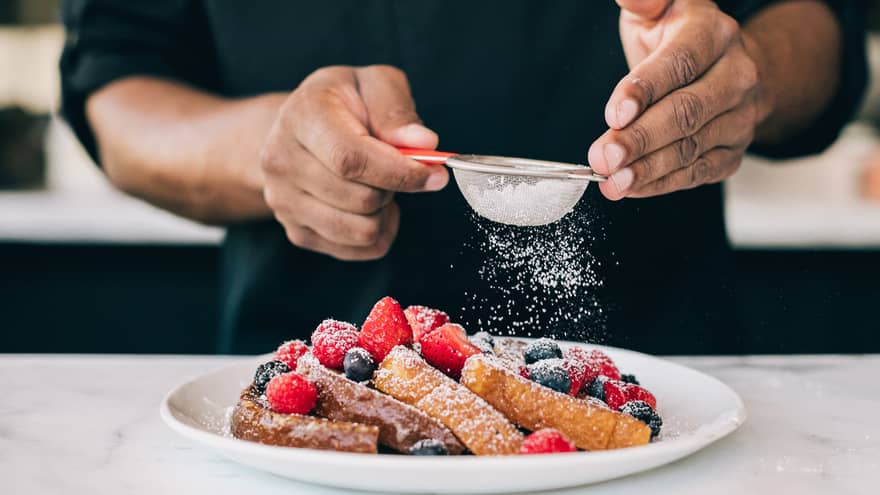 Four slices of French toast topped with raspberries, blueberries and strawberries being sprinkled with icing sugar through small sieve, on white plate on top of white marble counter