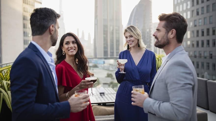 Four people dressed in cocktail attire stand talking and holding drinks on a rooftop terrace with a city skyline in the background