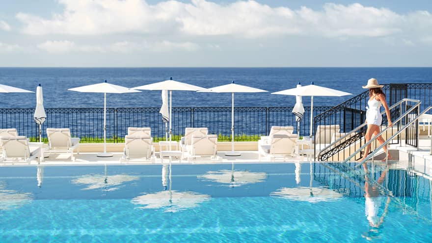 Woman in white with straw hat walks down steps into chaise-lined pool by Mediterranean