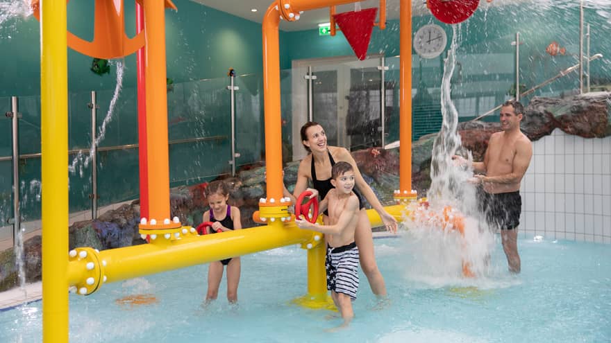 A man, woman and two children play in an indoor family pool with fun water pumps and spouts