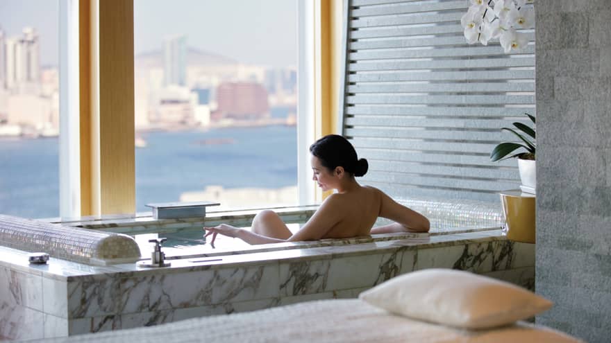 Woman's bare back above marble spa tub in tile bathroom by window with water, city views