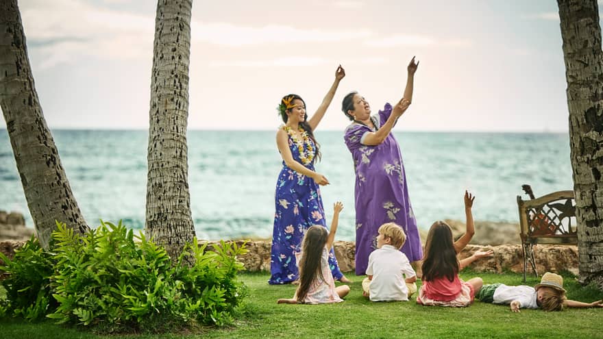 Young children sit on lawn by ocean, watch two Hawaiian women demonstrate dances
