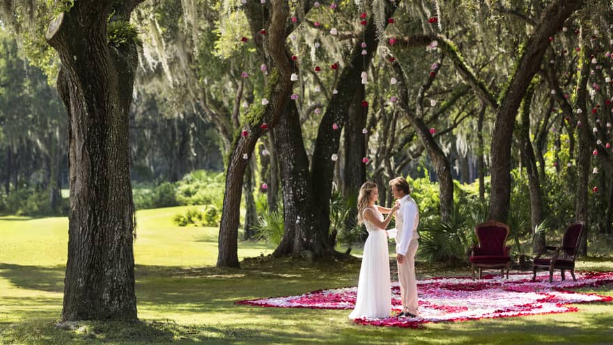 A whimsical outdoor wedding ceremony at Four Seasons Resort Orlando. The couple stands on a vibrant red rose petal carpet, exchanging vows beneath the enchanting Spanish moss-draped oak trees. A chic seating area with plush red velvet chairs awaits on the side, ready for an intimate celebration.