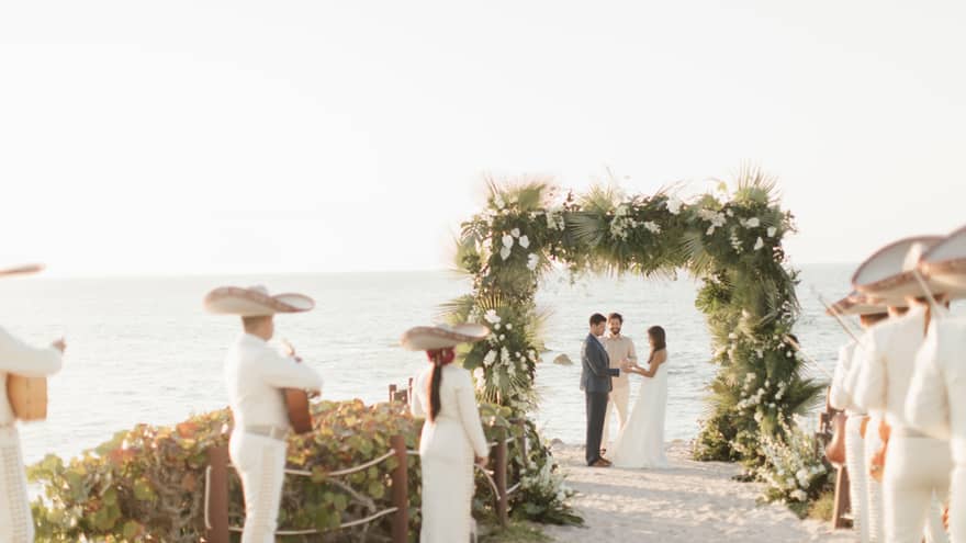 Bride and groom exchanging vows under a floral arch at Punta Mita cliffside wedding venue with ocean view, while a Mexican band plays music