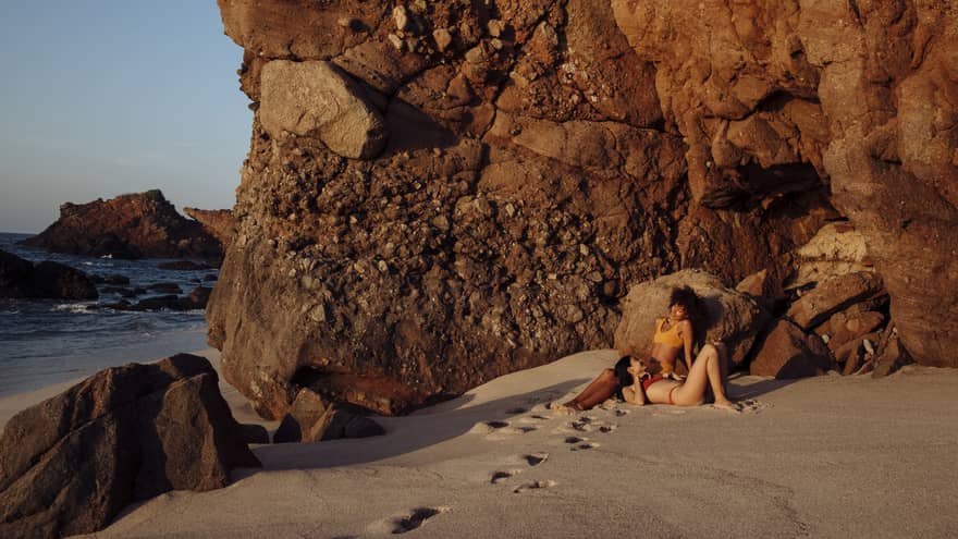 A woman is resting her head on another woman's leg as they relax on a beach near a rocky cliff.