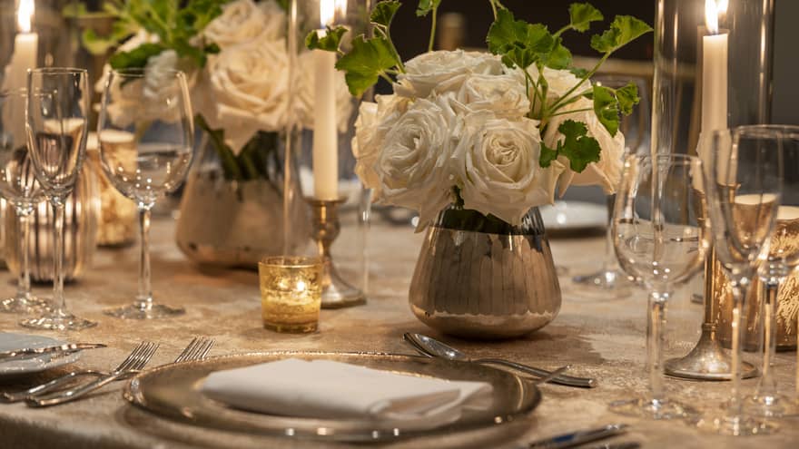 A dining table with white flower center pieces, silver plates, and wine glasses.