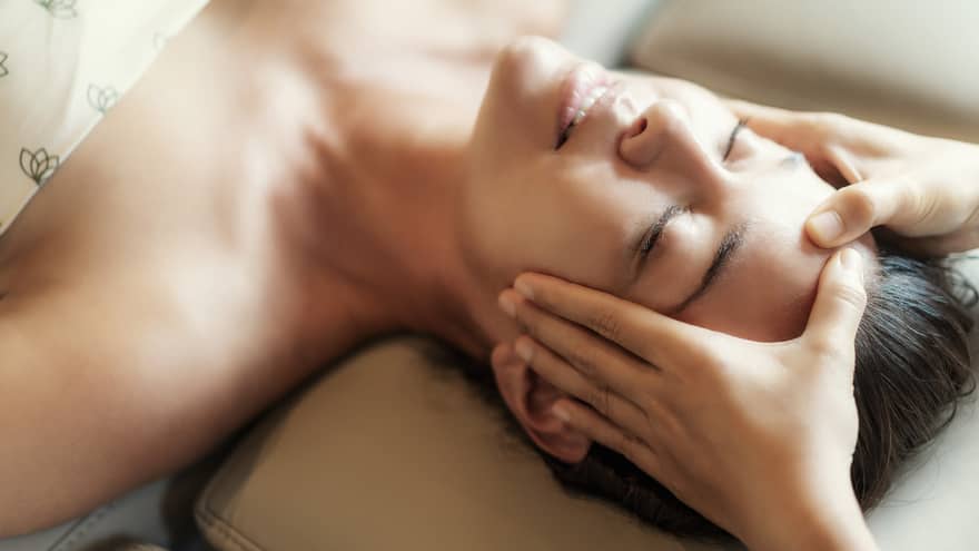 Spa facial, two hands rest on woman's forehead as she closes her eyes, lays under sheet on treatment table