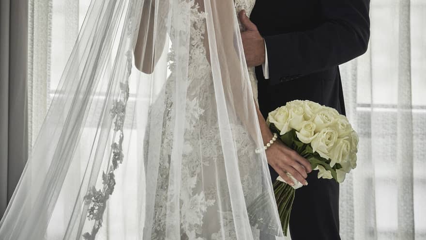 A wedding couple poses in front of a large window, dressed in wedding attire, with a long lace veil, holding a bouquet of white roses