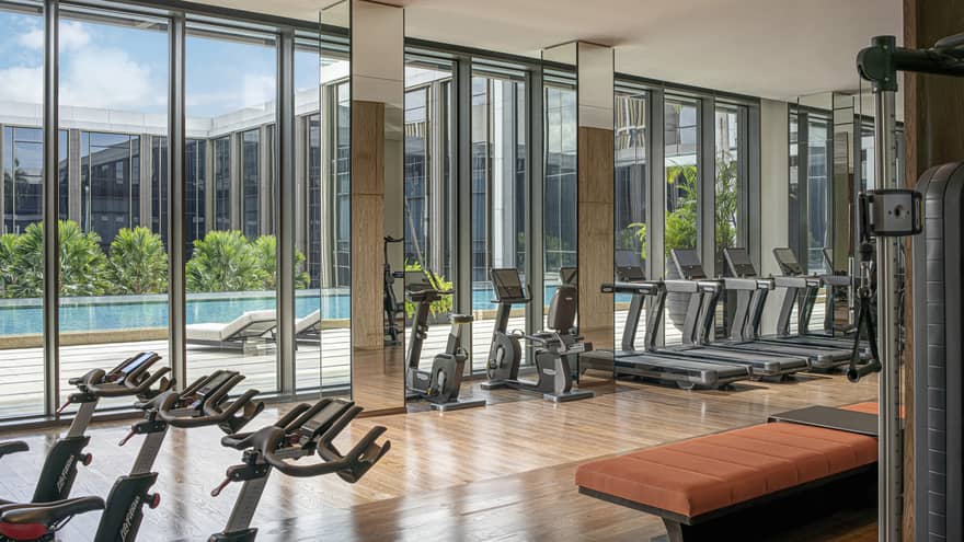 Hotel gym with floor-to-ceiling windows and rows of exercise equipment, natural light, pool views