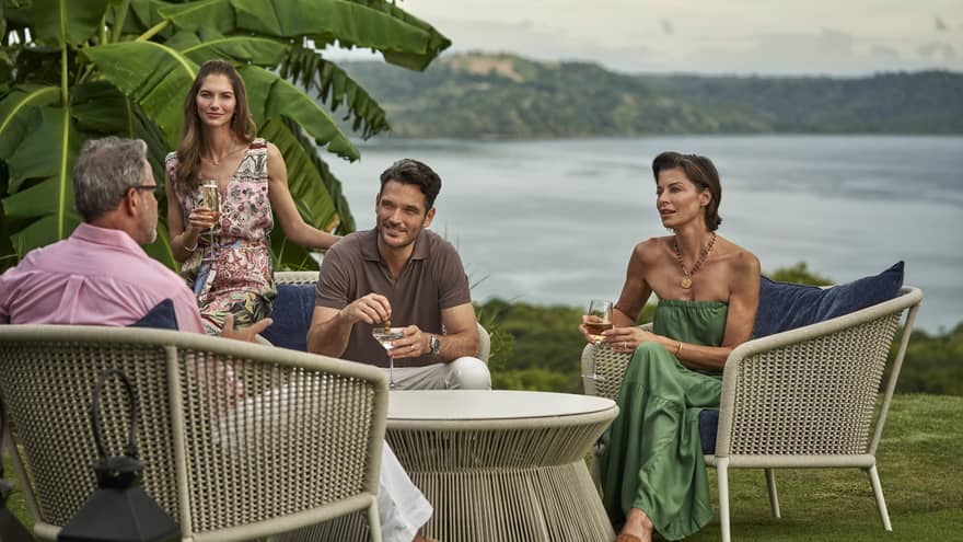 Four people sit around an outdoor table on a lawn overlooking the ocean, each with a drink in hand