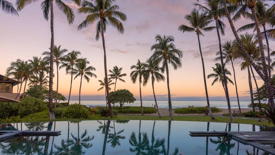 Luxury outdoor resort pool with view of ocean at sunset, surrounded by palm trees, at Four Seasons Resort Hualalai, Hawaii