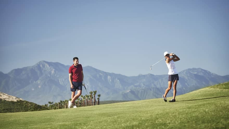 Woman swings golf club as man watches on green course, mountains in background