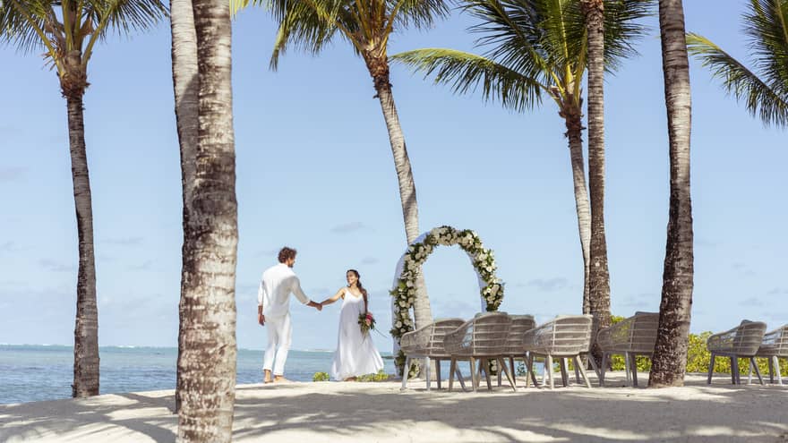 Newlyweds hold hands next to a flower arch on the beach