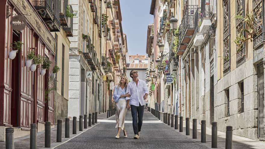A couple strolls hand in hand down a charming narrow street lined with colourful buildings and balconies, enjoying a sunny day in the city.