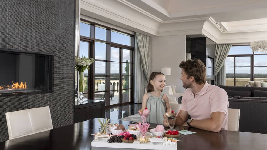 Man and young girl sit at dining table in hotel suite, next to fireplace