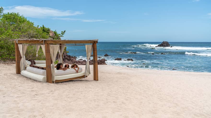 A cabana with three children laying on a plush seat underneath it on the beach, the ocean and plants are in the background.