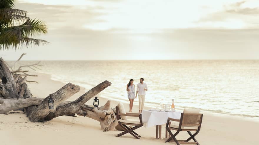 Couple walk across beach to small private dining table in sand at sunset