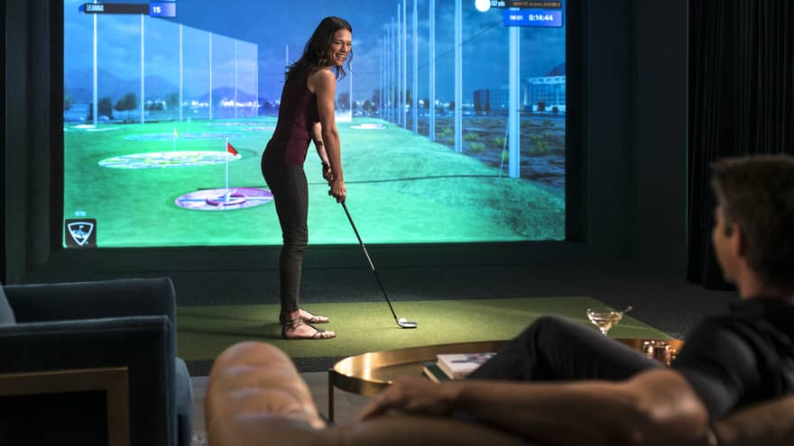 A woman prepares to swing a golf club in front of a virtual golf simulator screen