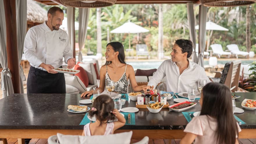 Chef serving a family seated at an outdoor dining table, with various dishes. The setting includes tropical décor, a poolside view and relaxed ambiance. 