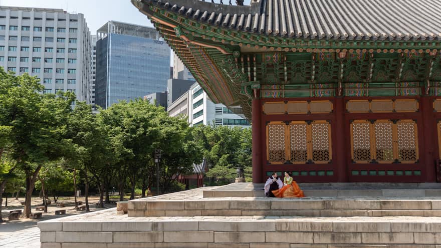 A couple in traditional Korean dress are seated on stone steps under the colourful roof awning of a tree-lined palace.