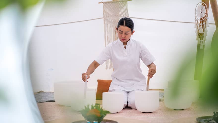 A person in white performs a healing ritual with crystal bowls inside a tranquil wellness tent, surrounded by soft décor and greenery