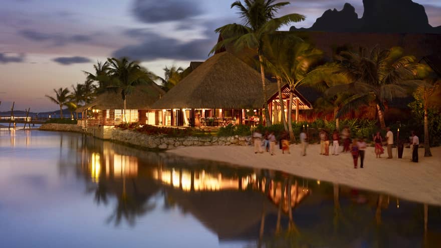 Guests on sand beach by pavilions with lights at night