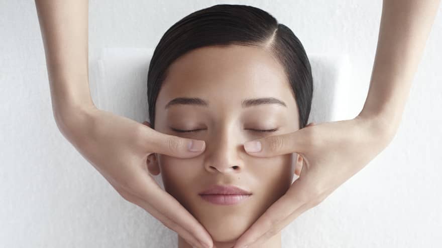 Woman in white bathrobe lies on massage table with eyes closed, masseuse rests hands on her cheeks