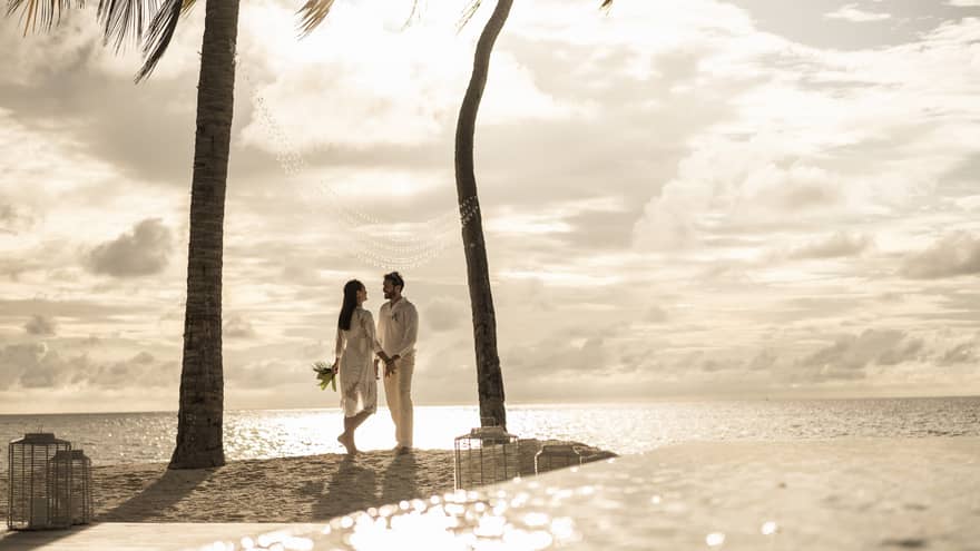 Couple stand between two palm trees at edge of sand beach at sunset