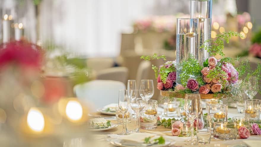 A wedding table setting featuring soft flickering candles in long glass holders, pink roses centerpiece and champagne glasses