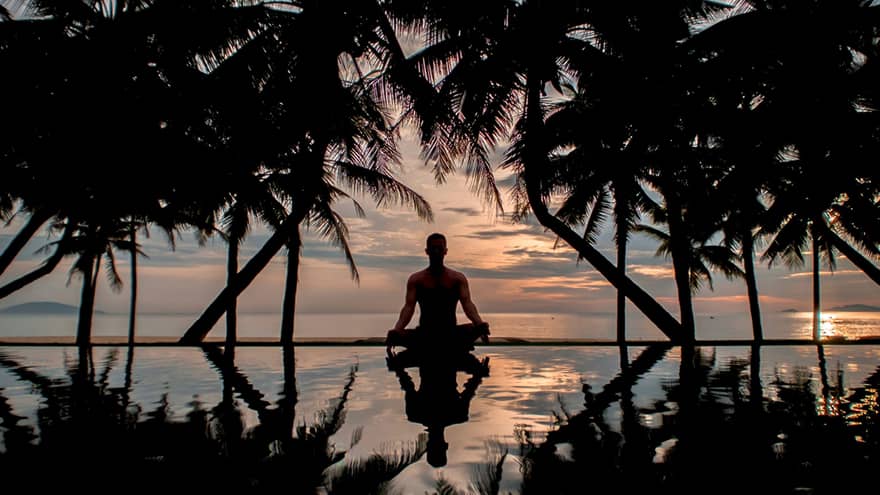 A man sitting in a yoga position at sunset next to an infinity pool surrounded by shadowy palm trees and vast ocean views