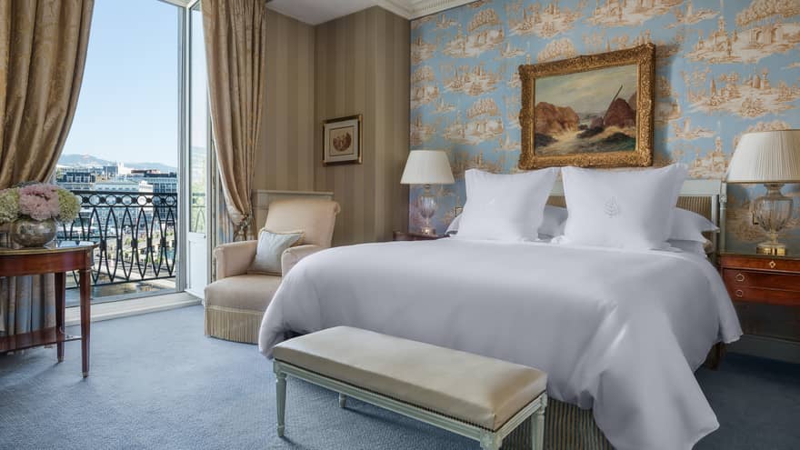 Luxury hotel room with a king bed, nightstands with lamps, bench, patterned wallpaper, armchair, framed artwork and a balcony with city view