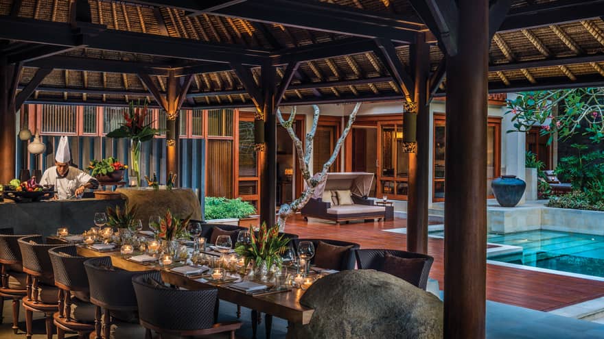 Chef prepares meal in front of elegant 10-person private dining table under thatched-roof gazebo, beside pool