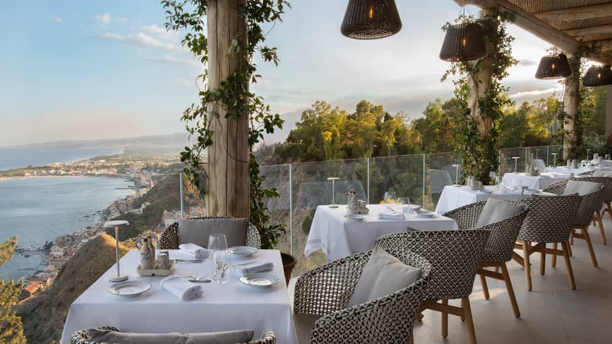 Outdoor restaurant terrace lined with 2-person tables, glass railing and sea view